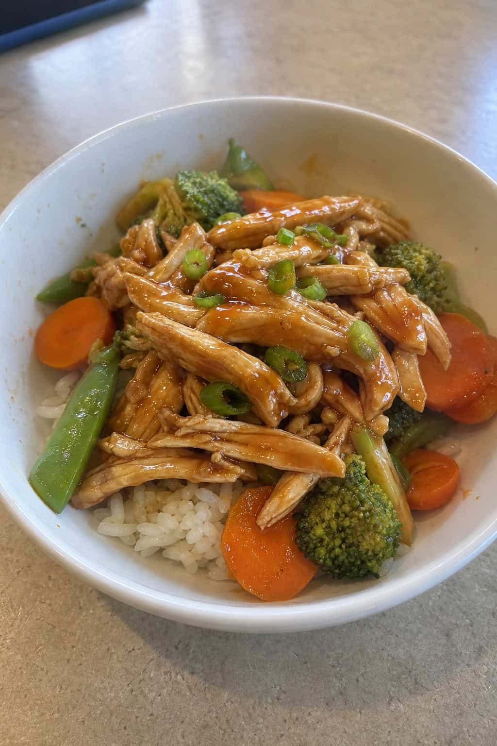 A white bowl of glossy honey-garlic shredded chicken served over steamed white rice with bright green broccoli florets and a sprinkle of sesame seeds and sliced green onions.