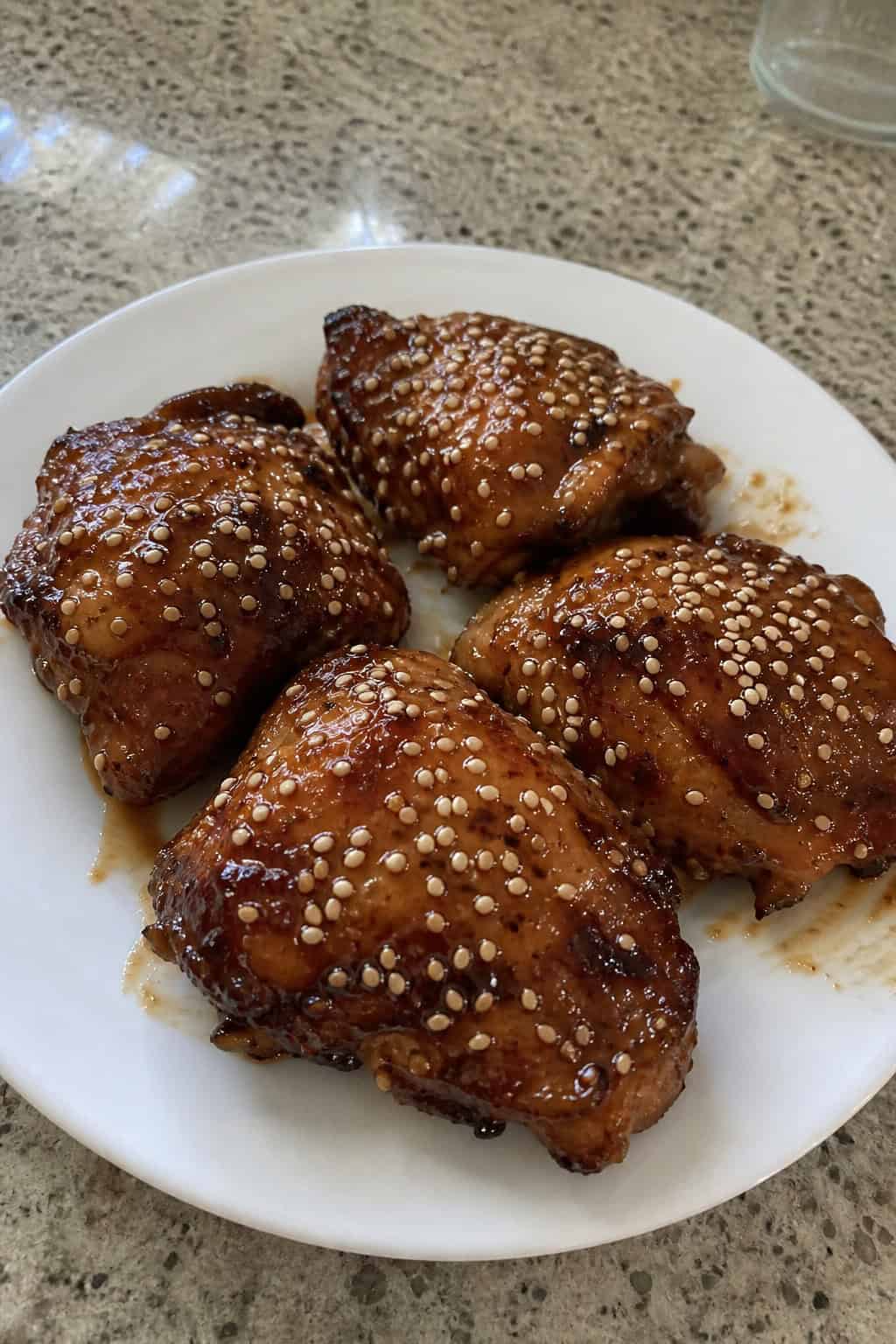 A plate of honey garlic soy-glazed chicken thighs topped with sesame seeds and garnished with fresh herbs on a wooden table.
