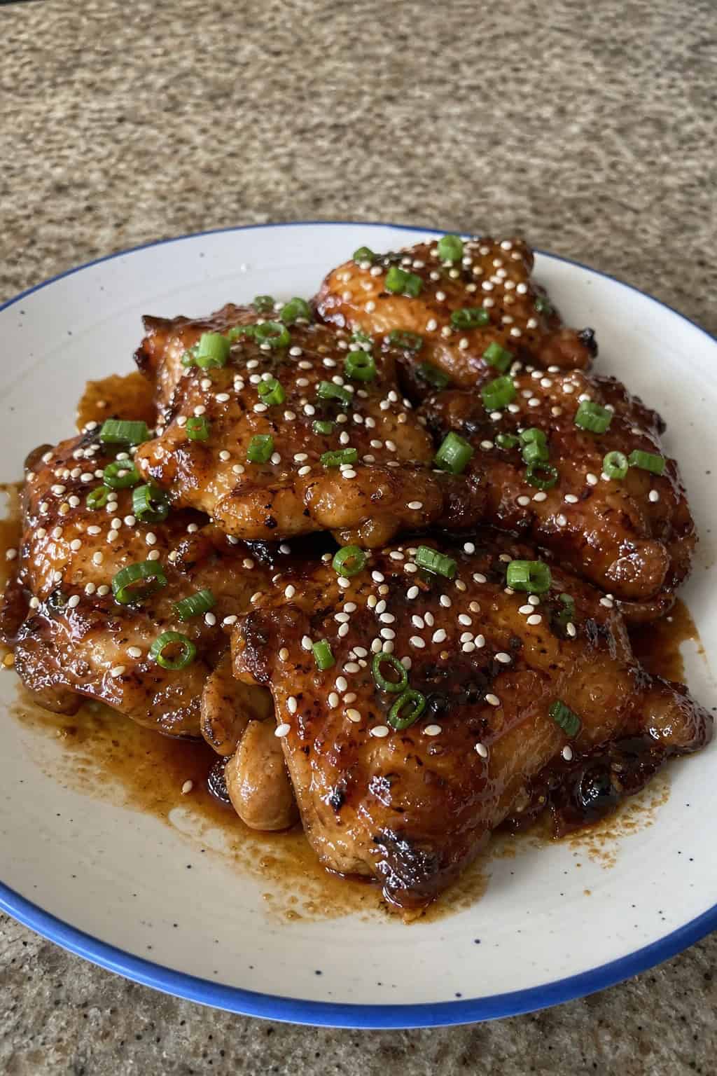 A plate of glazed honey garlic teriyaki chicken thighs garnished with green onions and sesame seeds on a white plate with garlic cloves and honey jar in the background.