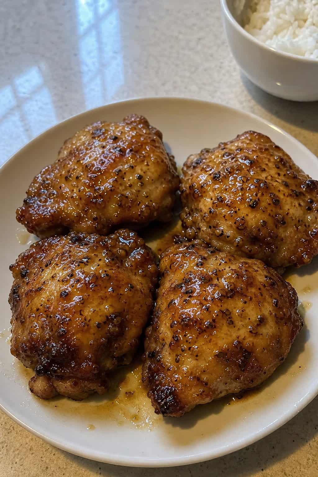 A plate of golden honey garlic yogurt-marinated chicken thighs garnished with fresh herbs, surrounded by small bowls of ingredients on a bright kitchen countertop.