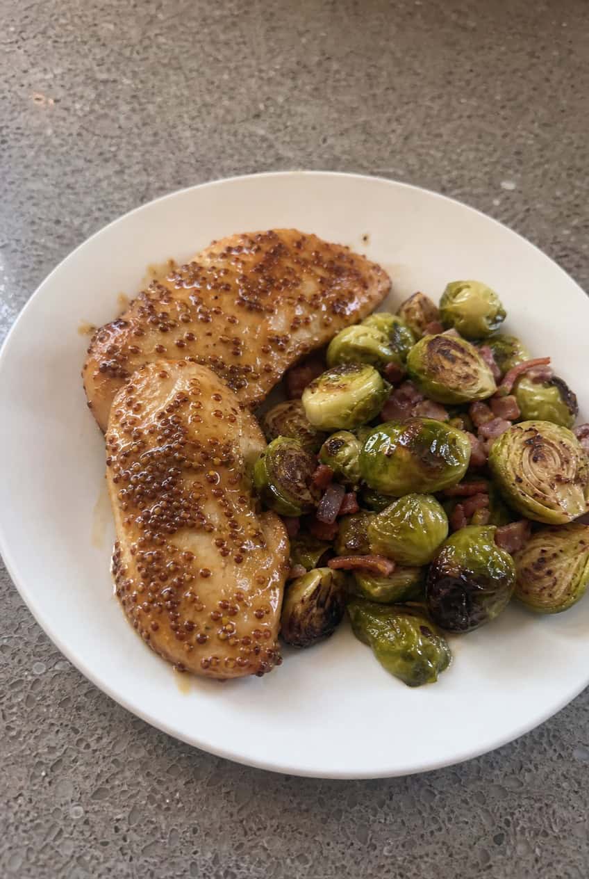 A white dinner plate holding honey mustard glazed shredded chicken alongside caramelized roasted Brussels sprouts with charred edges, served on a wooden table.