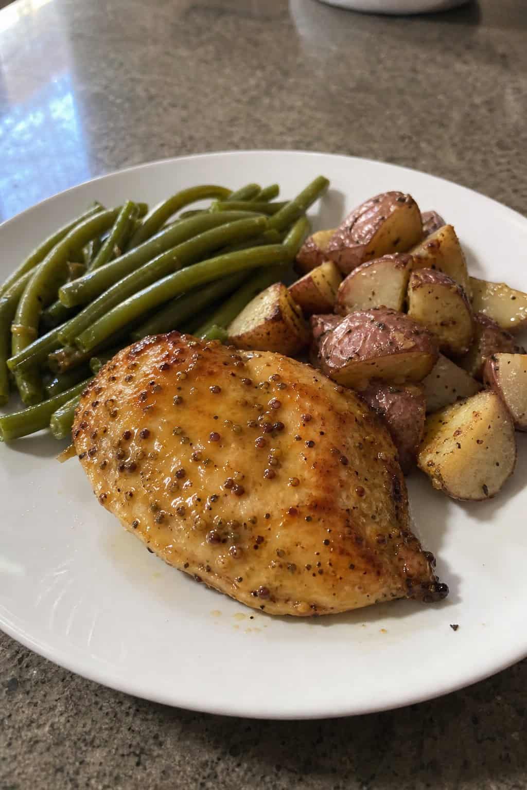 A plate with honey mustard chicken breasts, green beans, and roasted potatoes on a kitchen table.