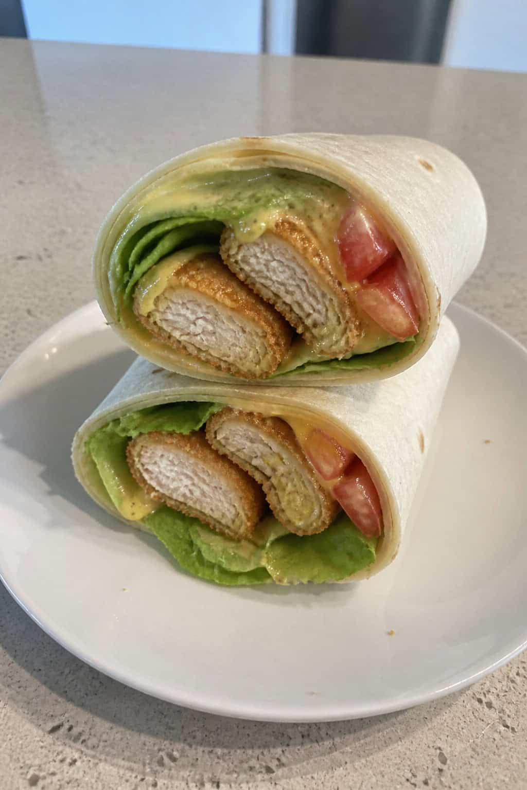 A honey-mustard chicken tenders wrap cut in half showing chicken tenders, lettuce, and tomatoes on a wooden table with small bowls of sauce nearby.