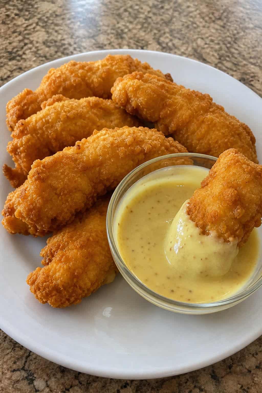 Close-up of golden chicken tenderloins on a wooden board with a small bowl of honey-mustard dipping sauce and scattered fresh herbs nearby.