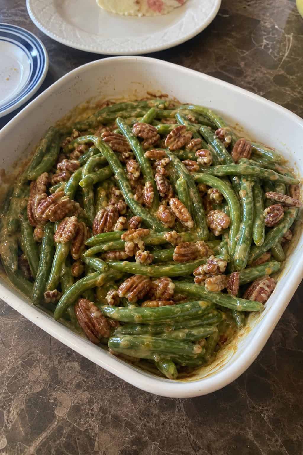 A honey-mustard green bean casserole topped with roughly chopped toasted pecans and a golden panko crust in a white ceramic baking dish on a wooden table.