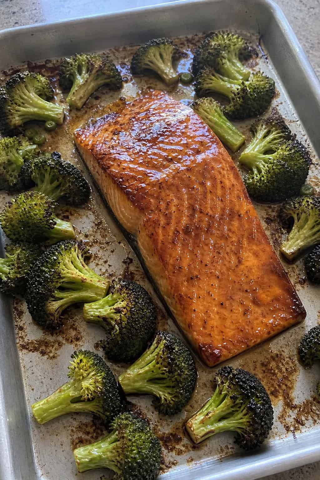 Skin-on salmon fillets glazed in a dark honey-soy sauce resting beside deeply charred broccoli florets on a foil-lined sheet pan.