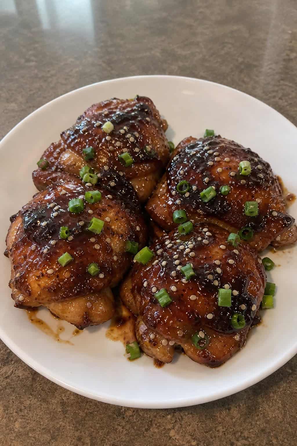 A plate of honey soy sticky chicken thighs garnished with sesame seeds and green onions on a wooden table with fresh ingredients around.