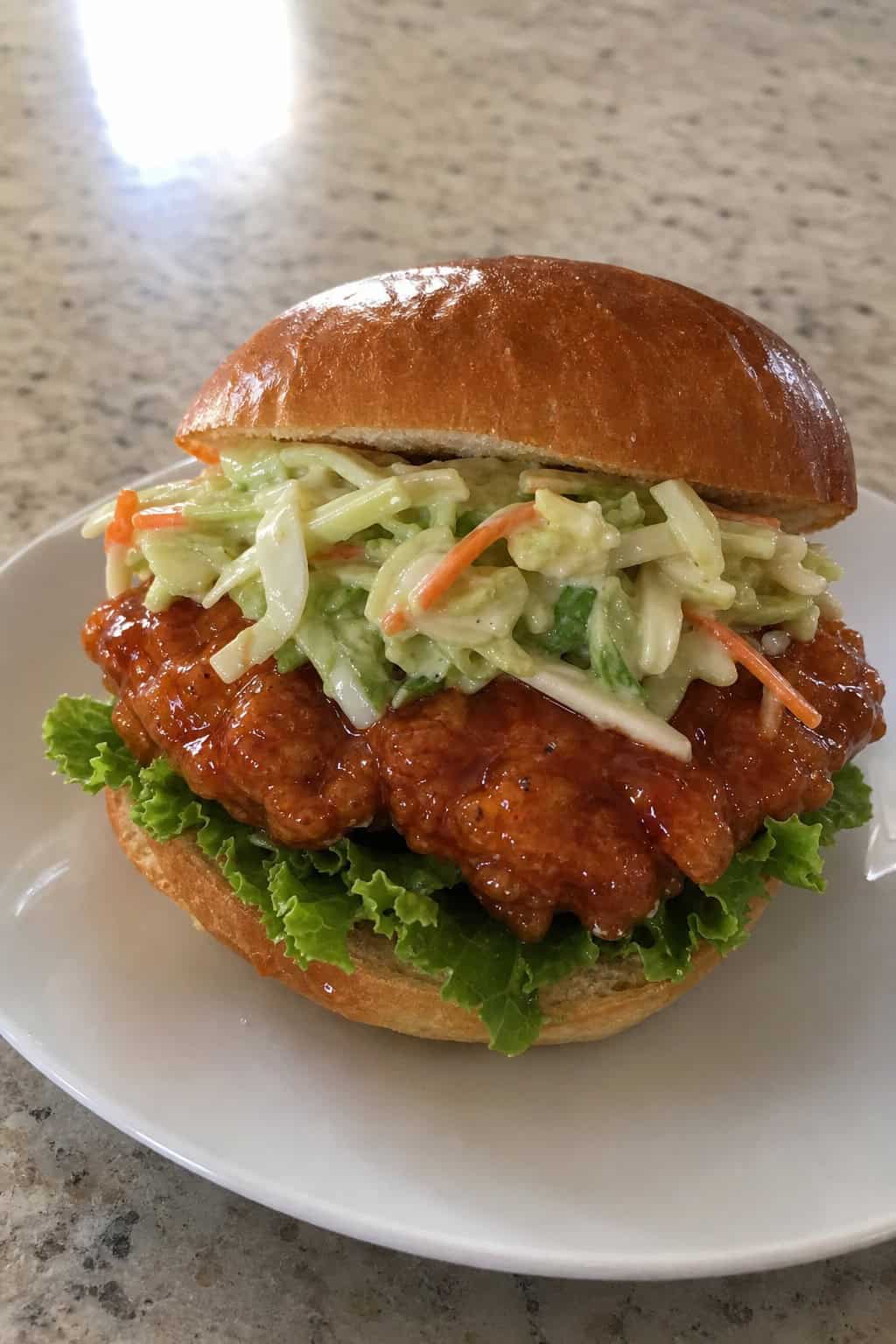 A honey-sriracha chicken sandwich with crispy fried chicken and lime slaw piled onto a toasted bun, served on a white plate