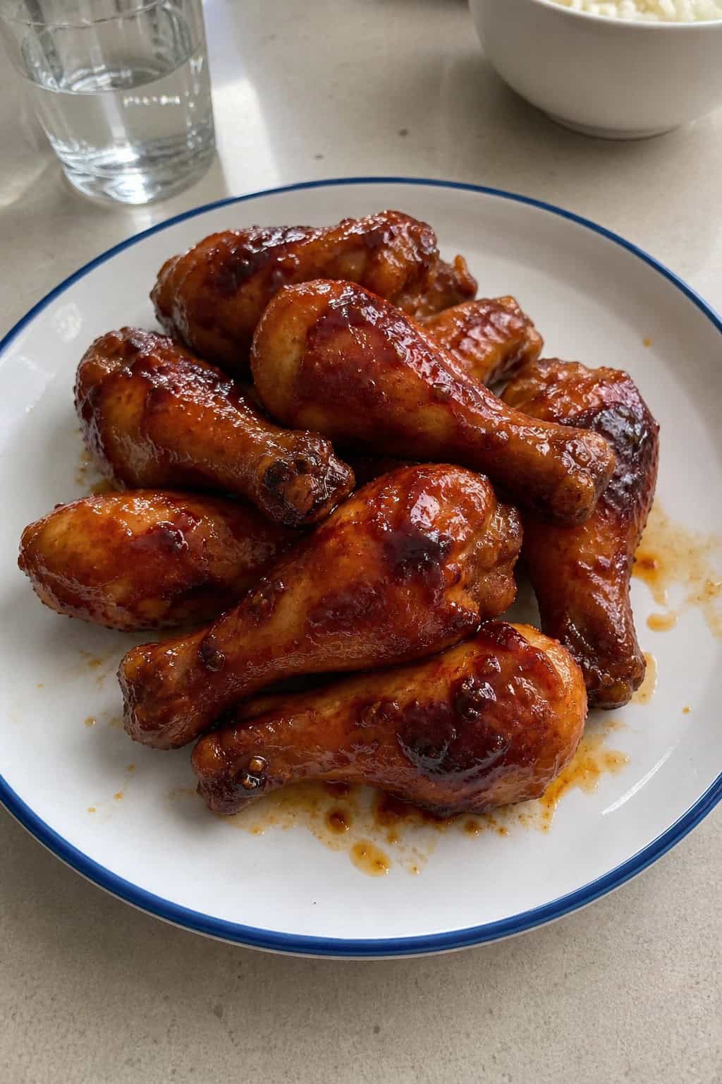 Deeply glazed honey-Sriracha chicken drumsticks on a white plate with bright herb garnish and two small sauce ramekins on a wood table.