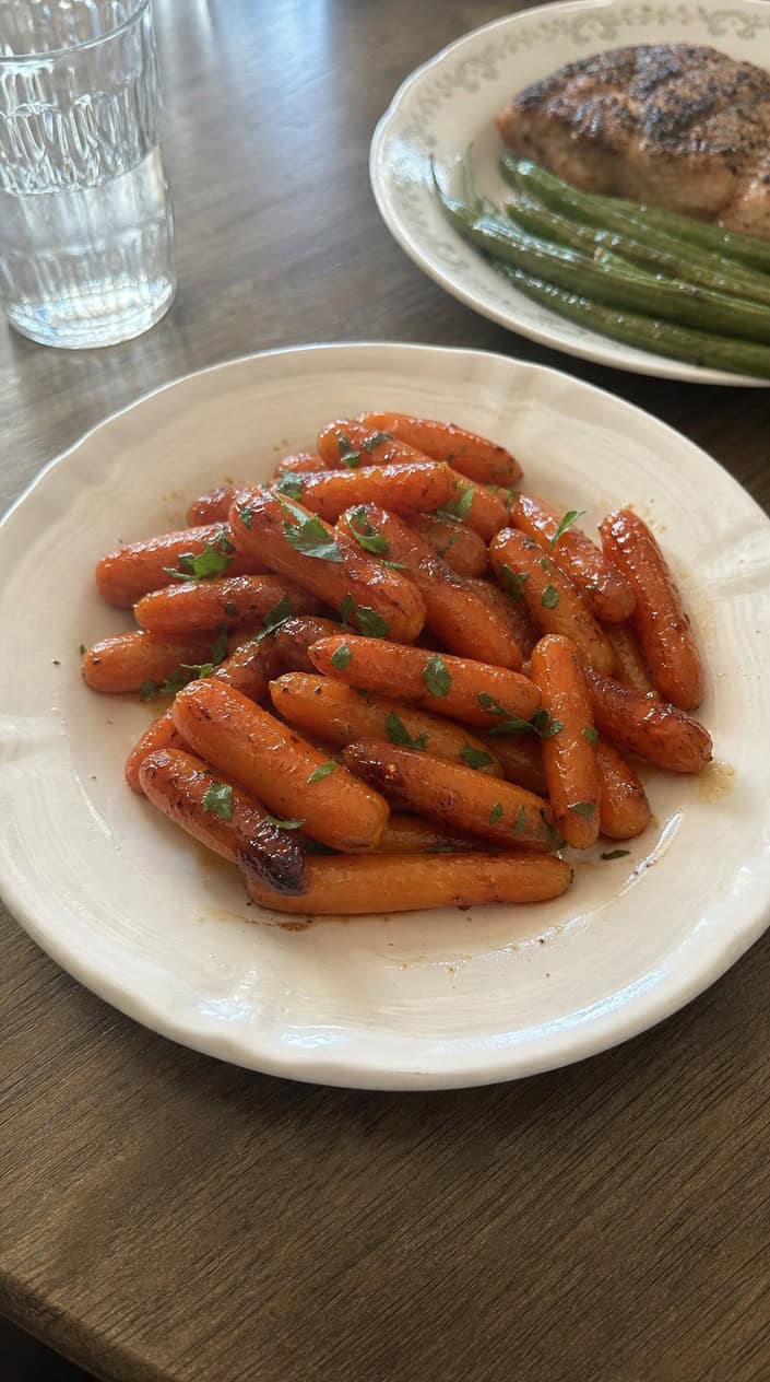 A plate of honey-sriracha glazed baby carrots garnished with fresh herbs on a wooden table.