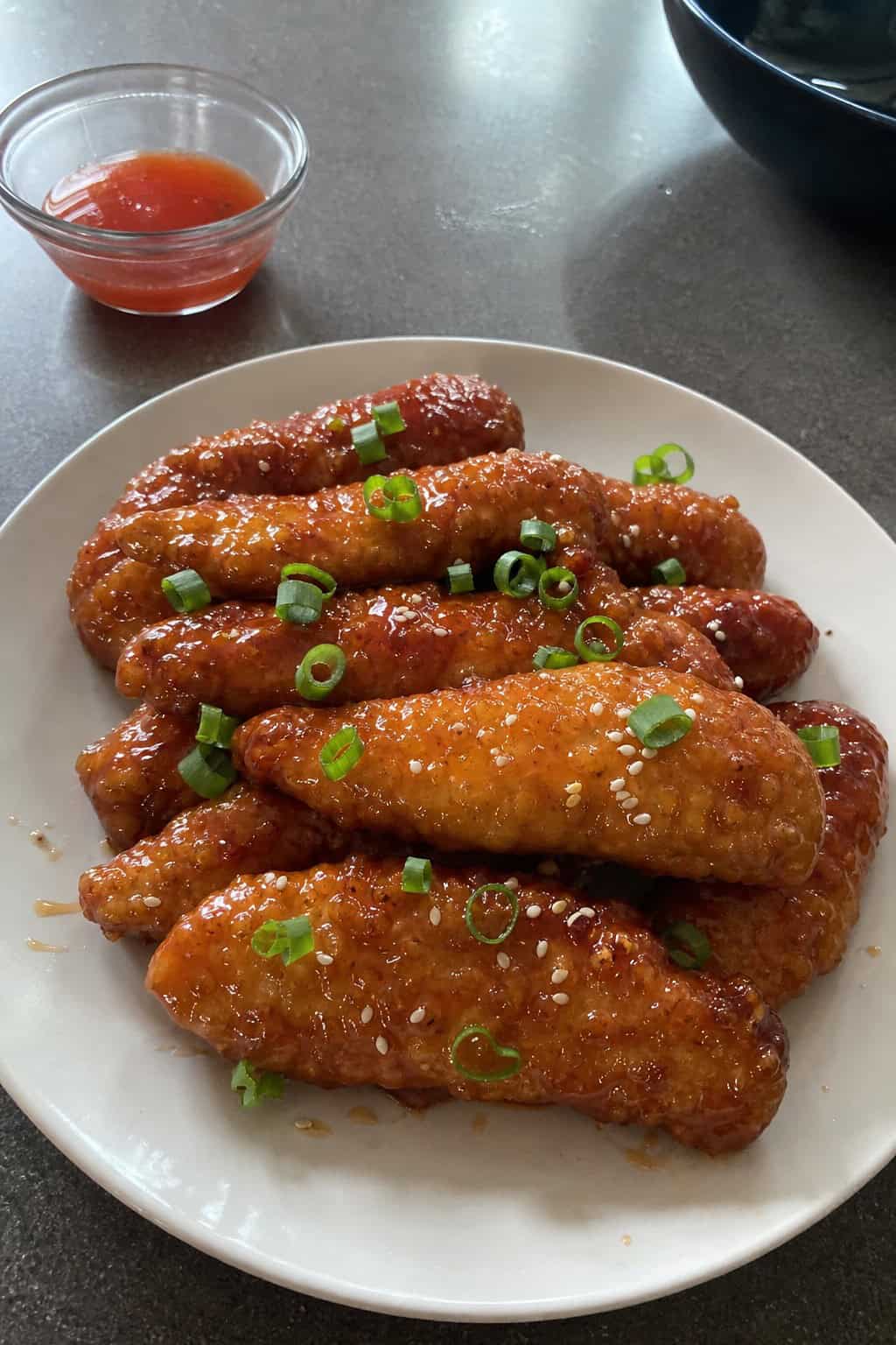 A plate of honey-sriracha glazed chicken tenders garnished with green onions and sesame seeds, served with dipping sauces and lime wedges on a wooden table.