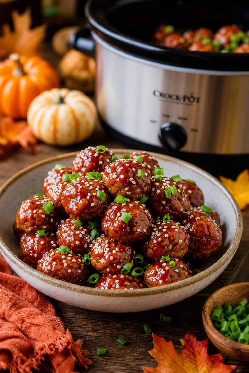 A bowl of glazed honey-sriracha meatballs garnished with sesame seeds and green onions on a wooden table with fall decorations and a slow cooker in the background.