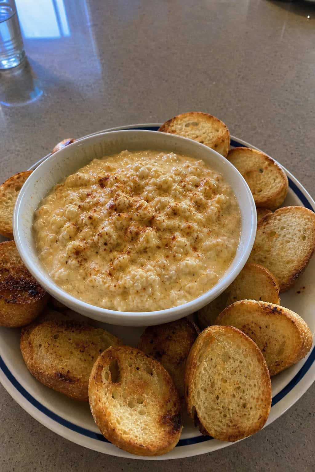 A bowl of hot crab dip garnished with seasoning and herbs, surrounded by toasted bread slices and vegetable sticks on a wooden board.