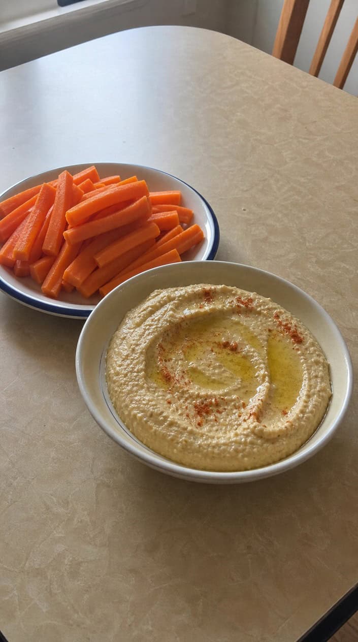 A bowl of hummus surrounded by fresh carrot sticks and other healthy vegetable snacks on a wooden table.