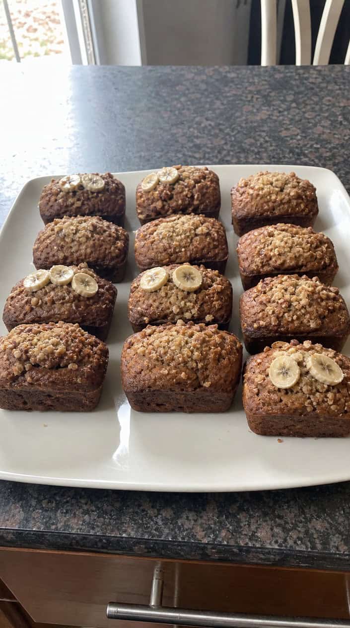 Six small golden-brown banana bread loaves arranged in two rows on a weathered wood surface, with ripe bananas, a cracked egg, and a small jar of flour visible in the background.