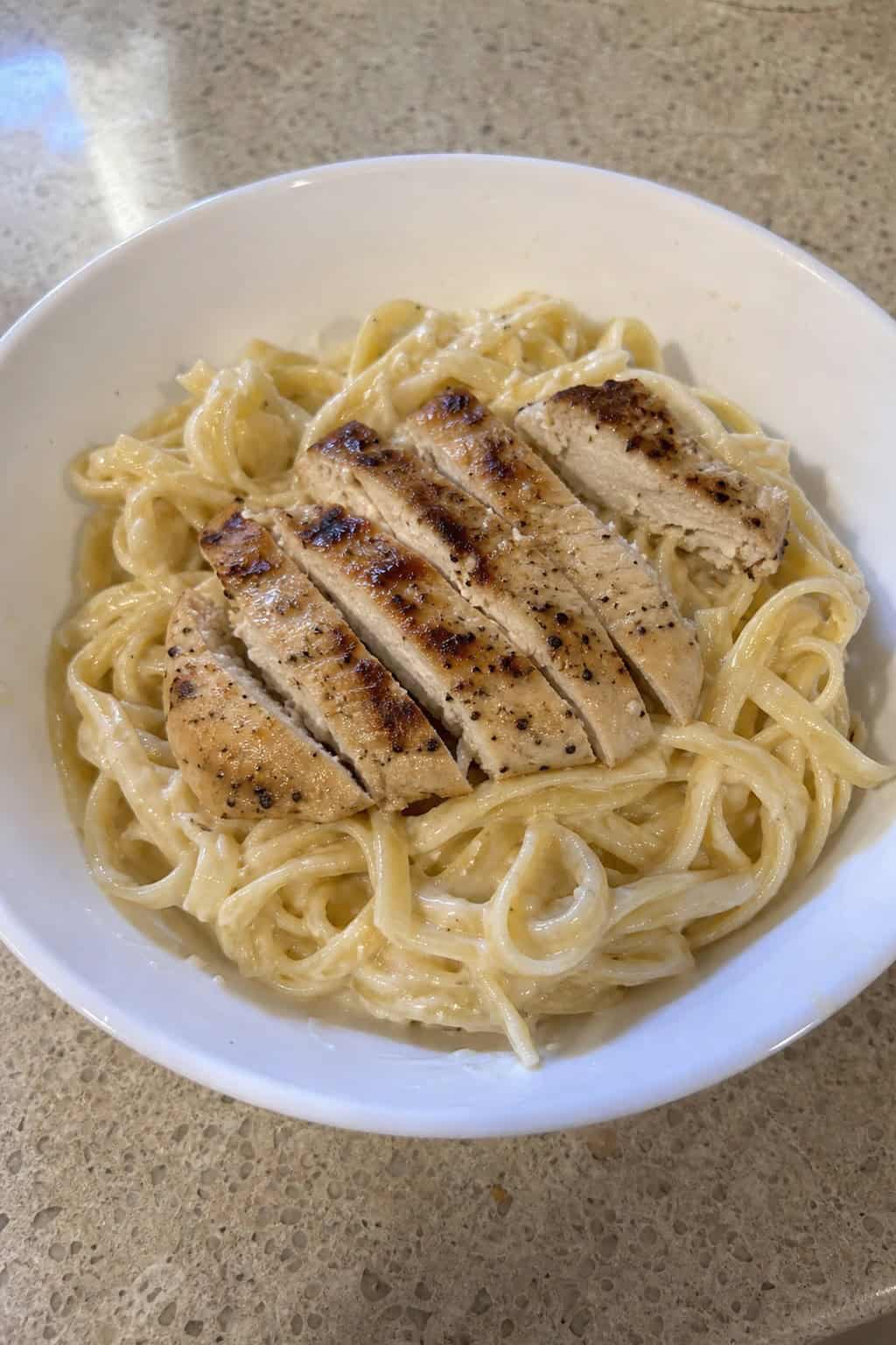 A bowl of creamy chicken Alfredo pasta garnished with parsley and Parmesan on a wooden table.