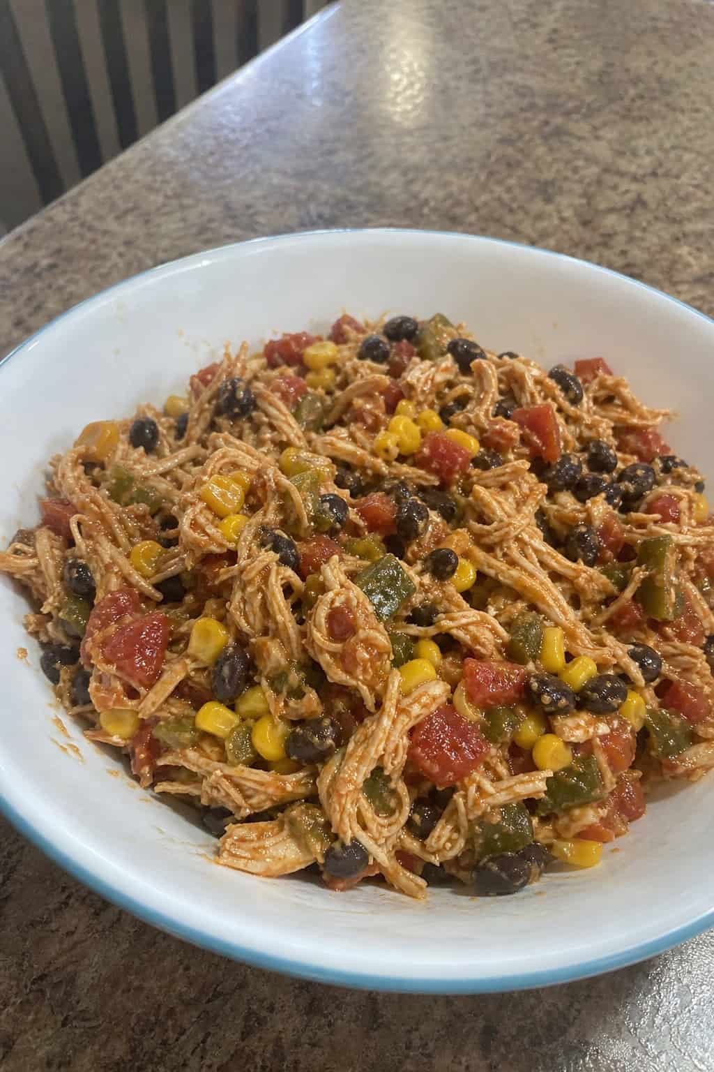 A bowl of shredded chicken enchilada filling with vegetables on a wooden table surrounded by fresh ingredients.