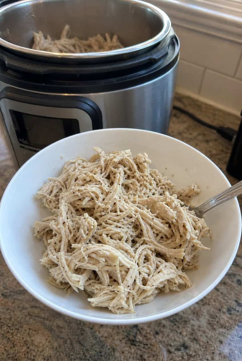 An Instant Pot with the lid off showing tender shredded chicken inside, surrounded by fresh garlic and herbs on a light kitchen countertop.