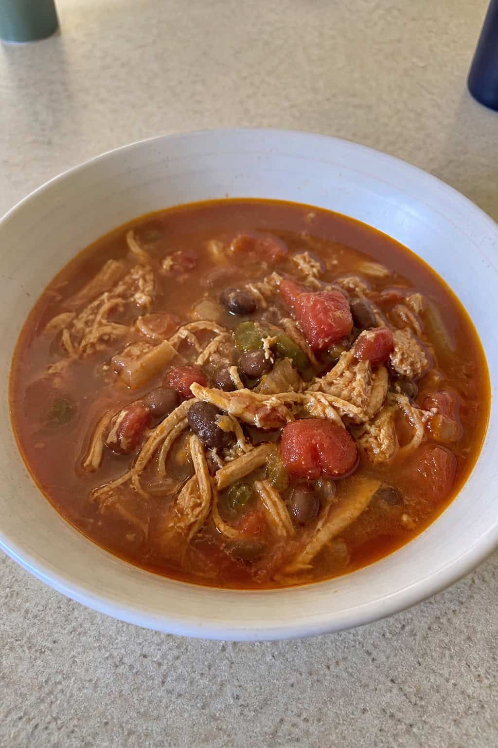 A deep bowl of shredded chicken chili with visible black beans and diced tomatoes, a spoon resting against the rim, surrounded by fresh cilantro sprigs on a wooden table.