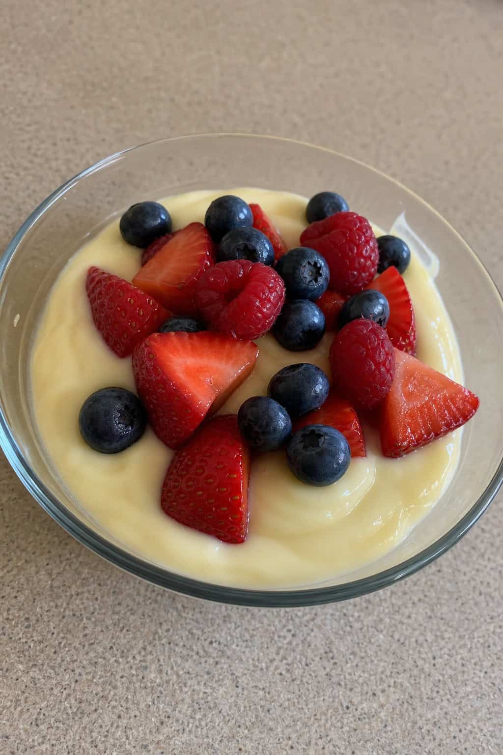 A bowl of vanilla pudding topped with fresh strawberries, blueberries, and raspberries on a wooden table.