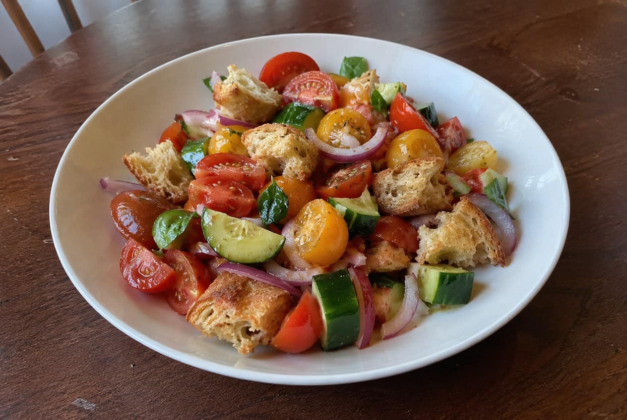 A rustic bowl of Italian panzanella with golden toasted bread cubes soaking in tomato juices, surrounded by chopped ripe tomatoes, sliced cucumber, thin red onion rings, and fresh basil, with whole tomatoes and a bottle of olive oil in the background.