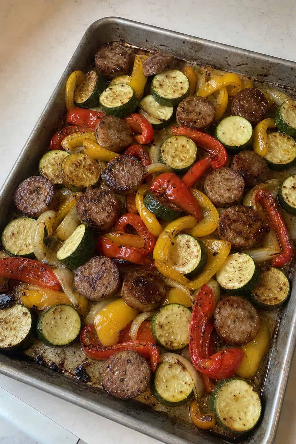 Sliced Italian sausage links roasted with thick zucchini rounds and red and yellow bell pepper strips on a foil-lined sheet pan set on a wooden table.
