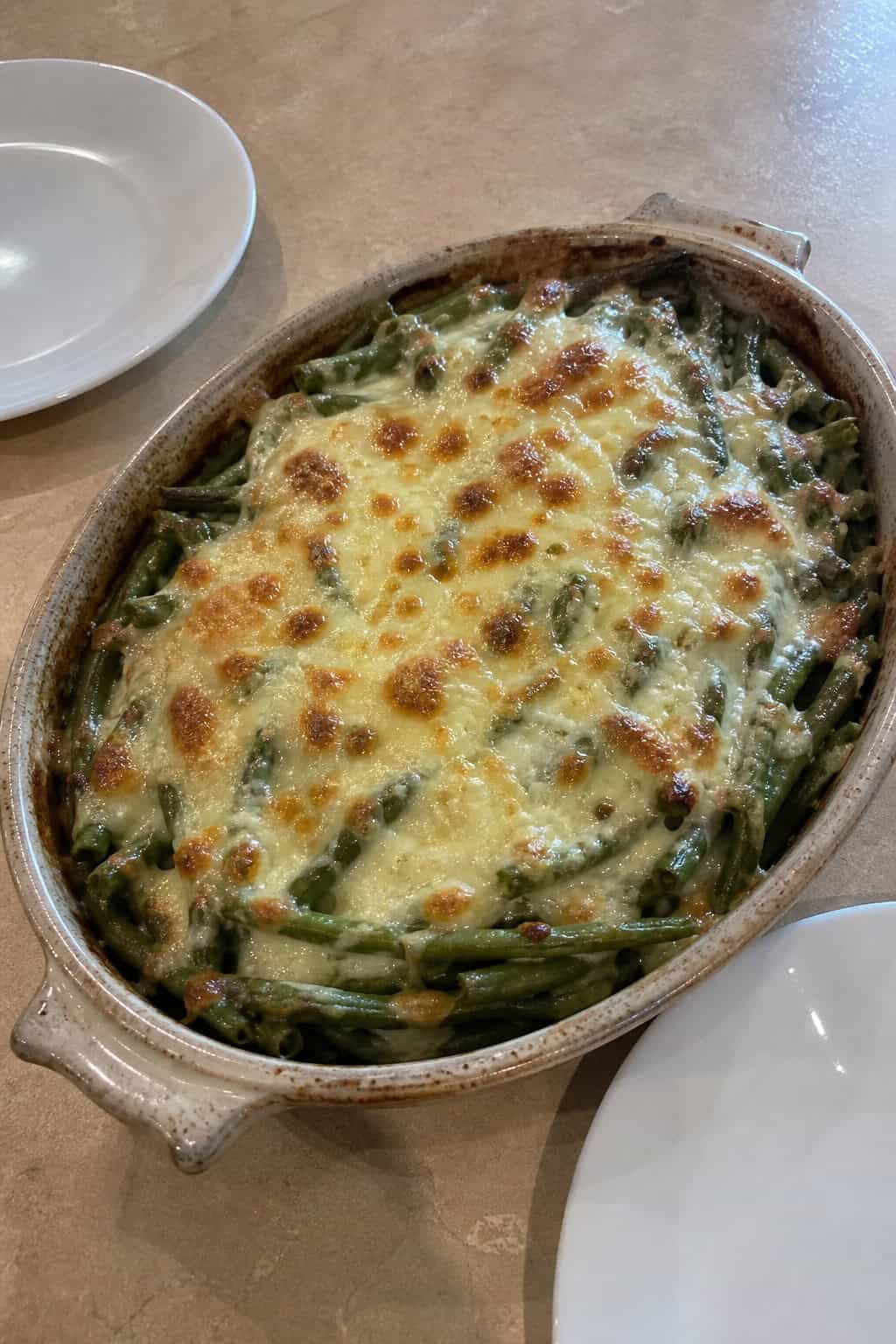 A baked green bean casserole topped with melted, lightly browned mozzarella and golden panko crumbs in a white ceramic dish, with fresh green beans and herb sprigs arranged on the wooden table around it.