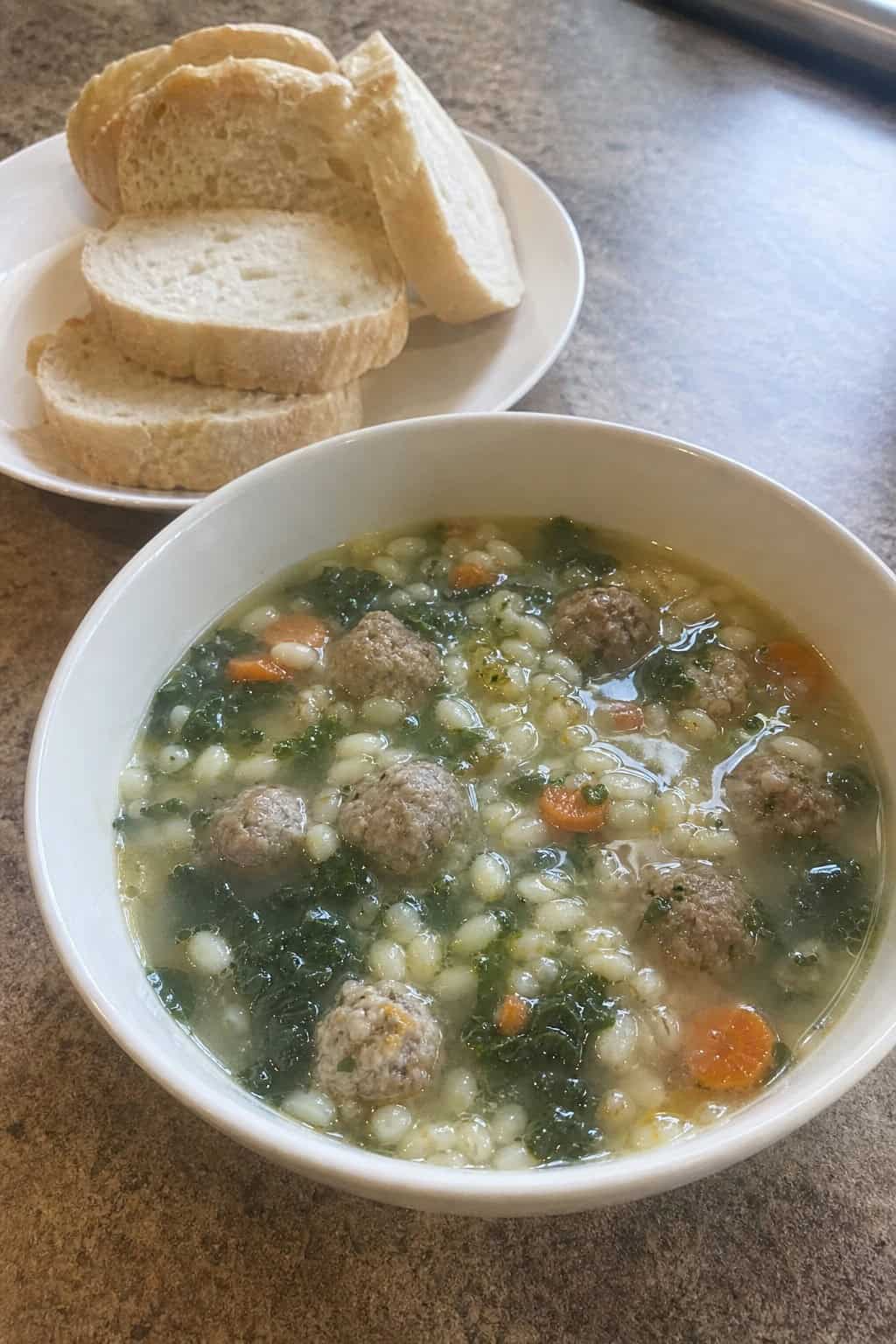 A bowl of Italian wedding soup with meatballs, greens, and pasta on a wooden table with fresh herbs and bread nearby.