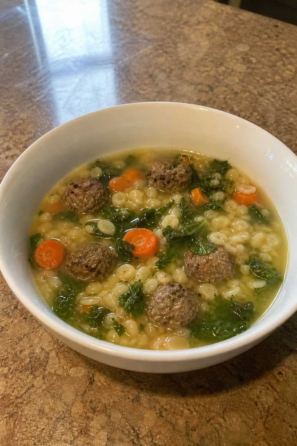 A bowl of Italian wedding soup with meatballs, pasta, and vegetables on a wooden table next to fresh herbs and a slow cooker.