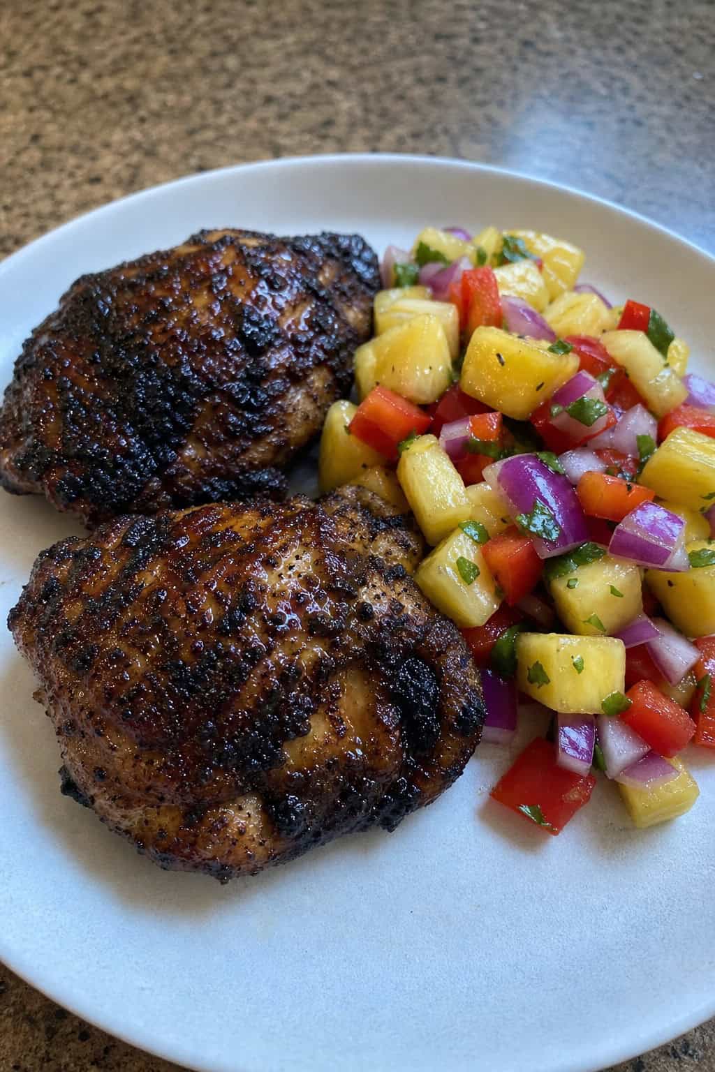 A plate of grilled Jamaican jerk chicken thighs served with pineapple salsa and garnished with lime wedges and fresh herbs on a wooden table.