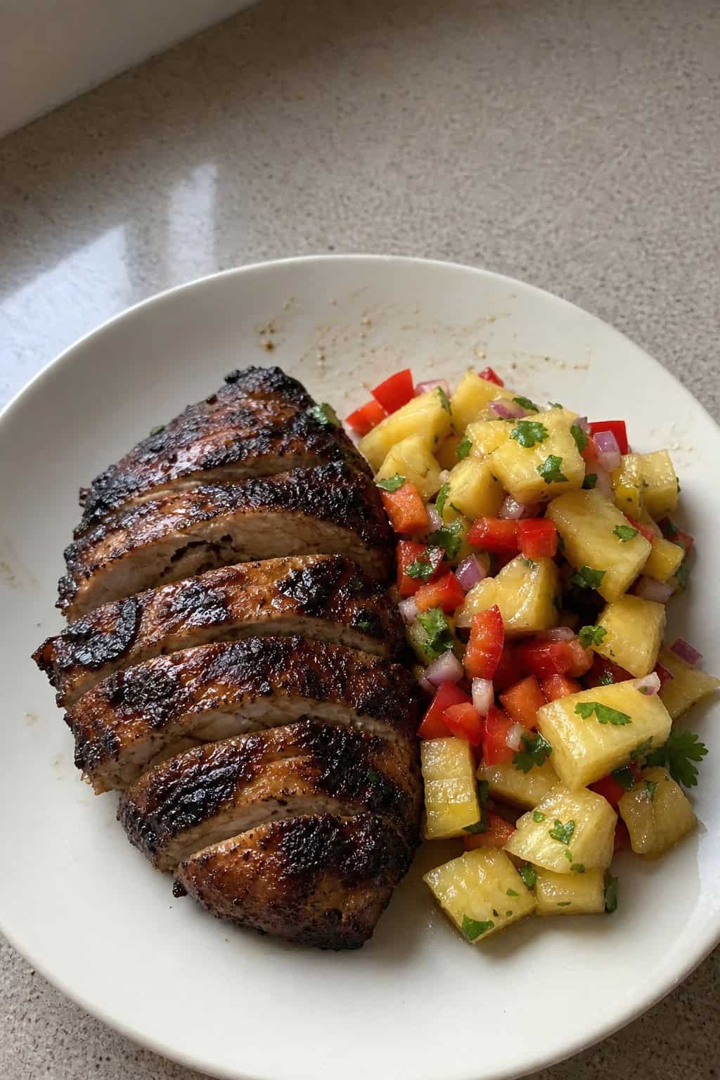 A grilled Jamaican jerk chicken breast served with pineapple salsa on a plate, garnished with lime wedges and fresh herbs.