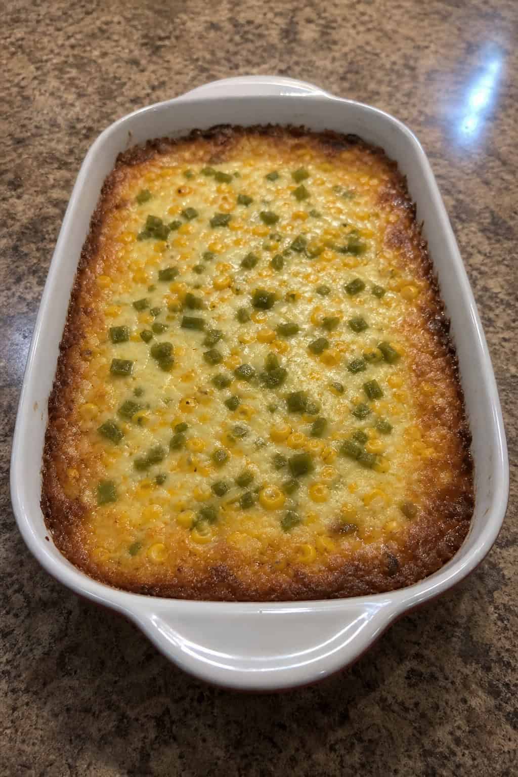 A golden baked corn casserole with visible green chile pieces under a layer of melted white Monterey Jack cheese in a ceramic dish on a wooden table.