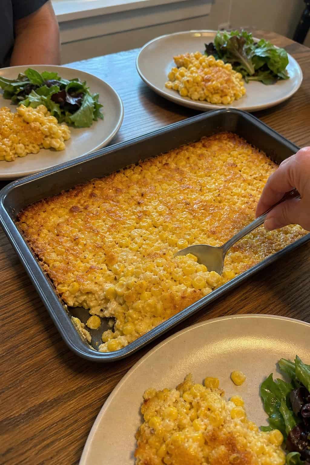 A large corn casserole in a clear glass baking dish on a wooden buffet table with several people in the background serving themselves onto white plates.