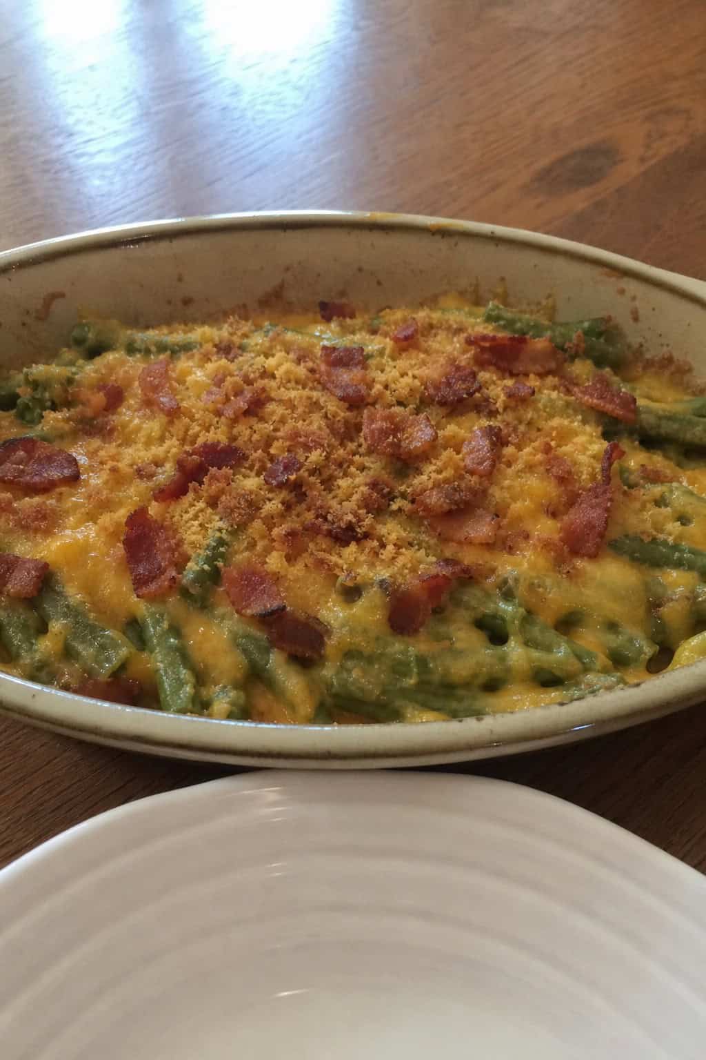 A green bean casserole topped with melted sharp cheddar and crispy bacon crumbles in a wide white ceramic dish on a wooden table.