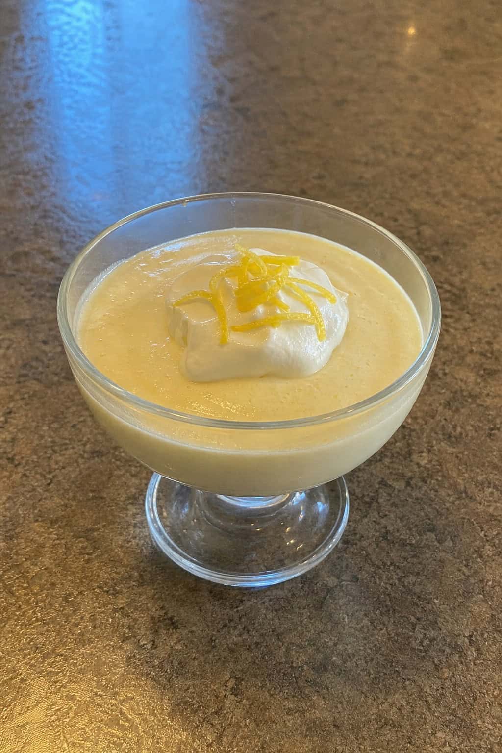A bowl of creamy lemon cheesecake pudding topped with mascarpone, lemon zest, and a mint leaf on a wooden table.