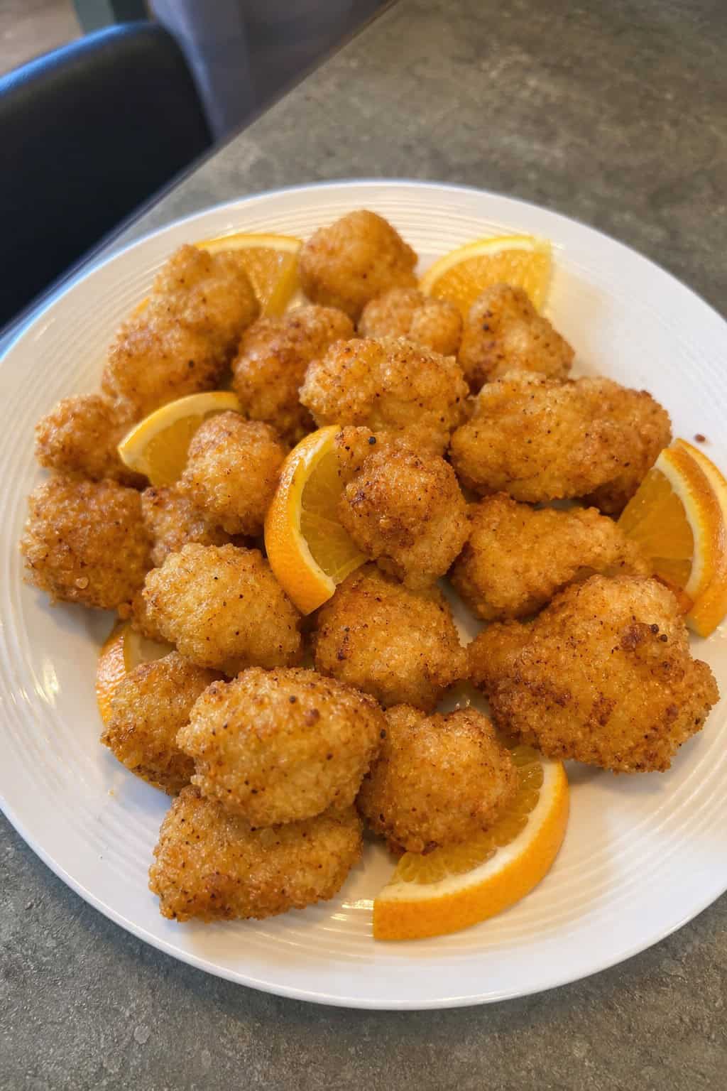A plate of small crispy citrus popcorn chicken bites with fresh orange slices and a bowl of dipping sauce on a kitchen table.