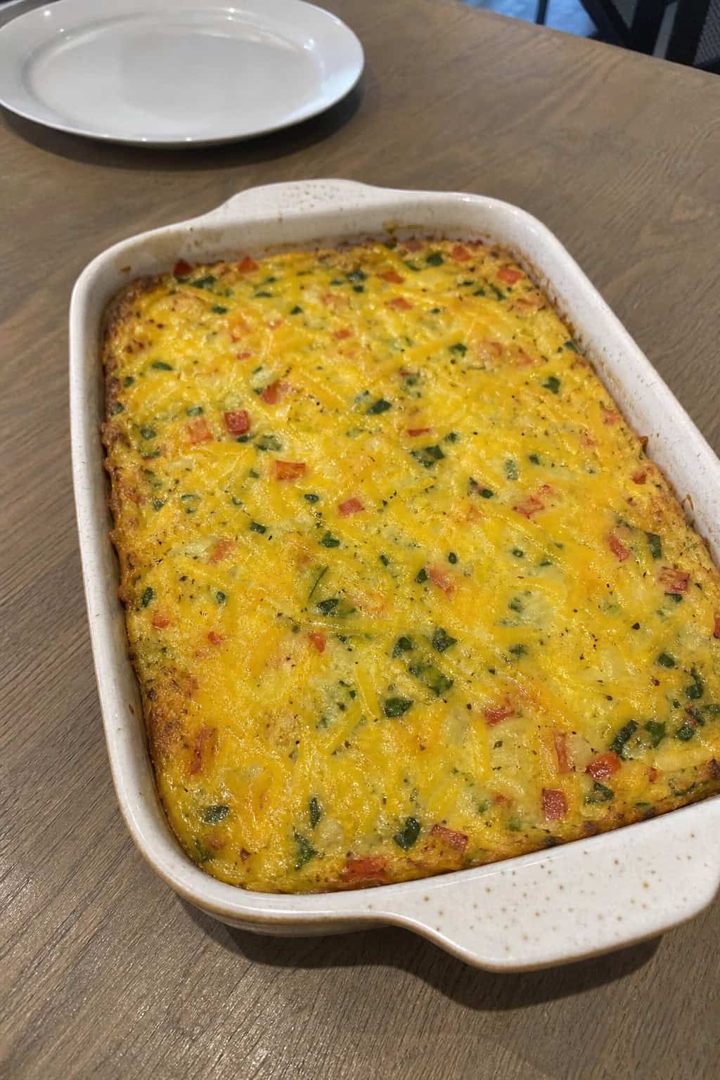 A golden hashbrown casserole in a ceramic dish with barely visible flecks of grated carrot and green spinach folded into the egg layer, on a wood table with a glass of orange juice beside it.