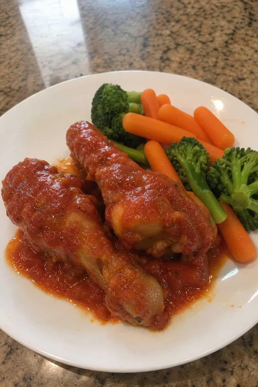 Chicken legs nestled in a rich red tomato sauce in a white baking dish, garnished with fresh basil leaves, with steamed vegetables on the side.
