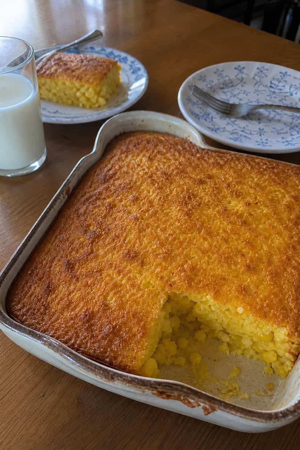 A pale golden sweet corn casserole in a round cream-colored ceramic dish on a wooden table with stacked white plates and simple silverware arranged beside it.