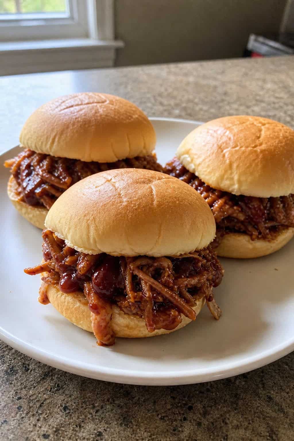 Close-up of pulled pork sliders on potato rolls arranged on a wooden board with herbs and BBQ sauce.