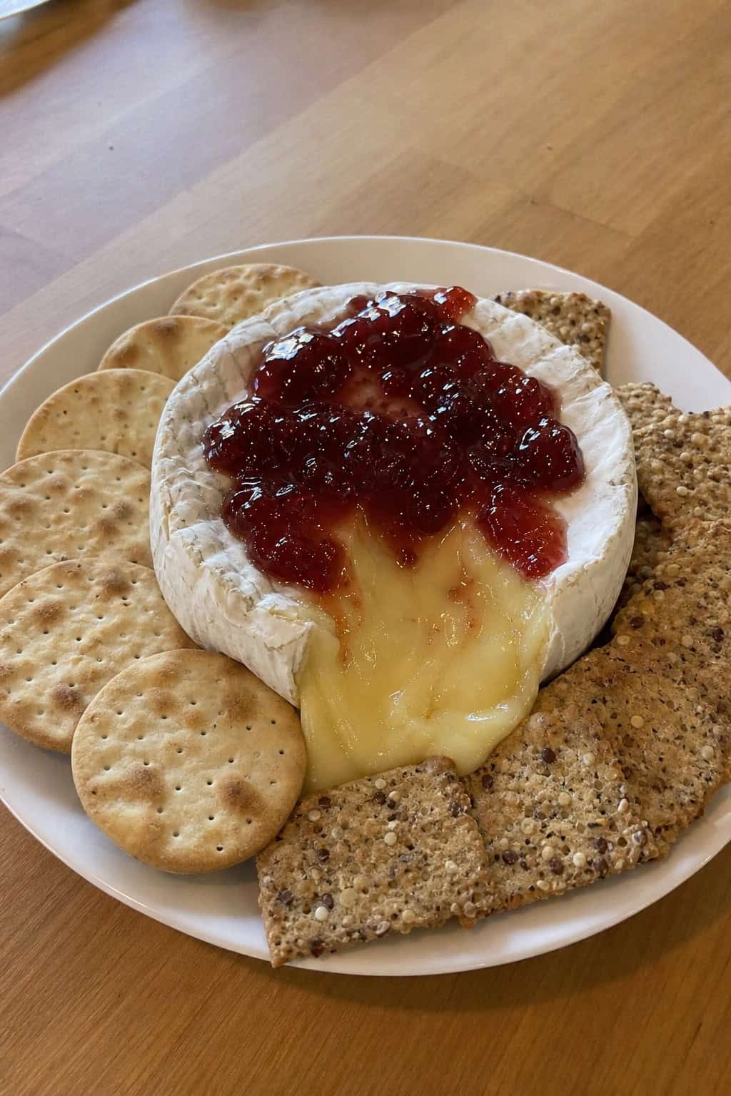 A warm Brie cheese wheel topped with jam on a wooden board surrounded by assorted crackers.
