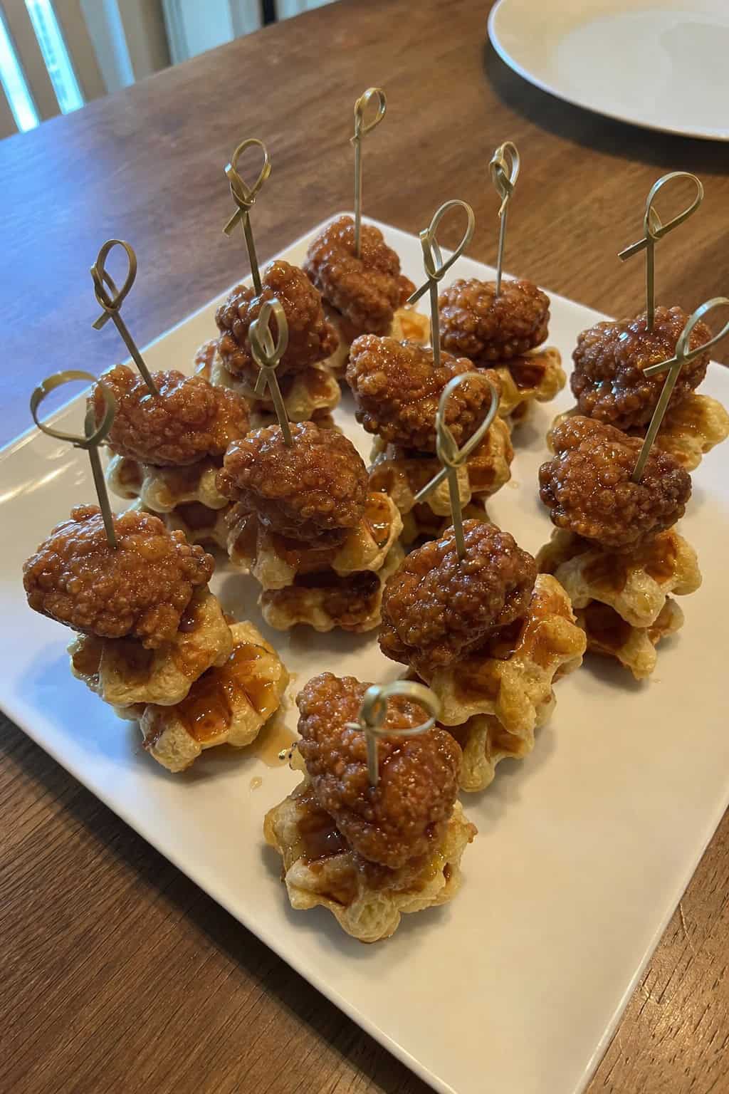Mini chicken and waffles skewers arranged on a serving platter on a wooden table with a casual party setting in the background.