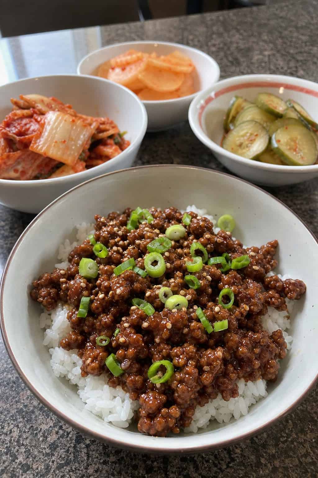 A bowl of Korean-style beef with rice, garnished with green onions and sesame seeds, surrounded by small side dishes on a wooden table.