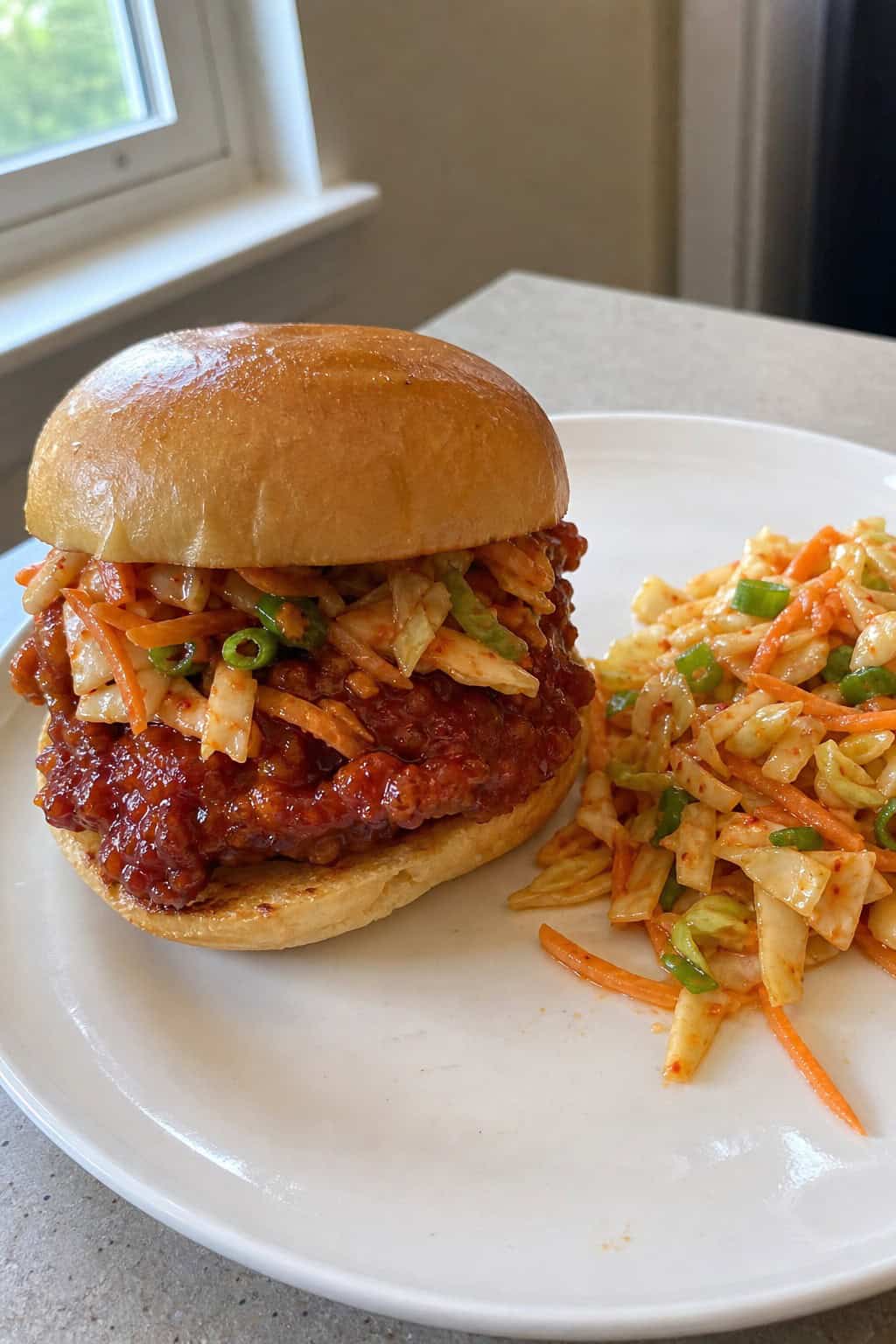 A spicy gochujang chicken sandwich with a vivid red sauce and colorful kimchi slaw piled onto a toasted bun, set on a wooden board