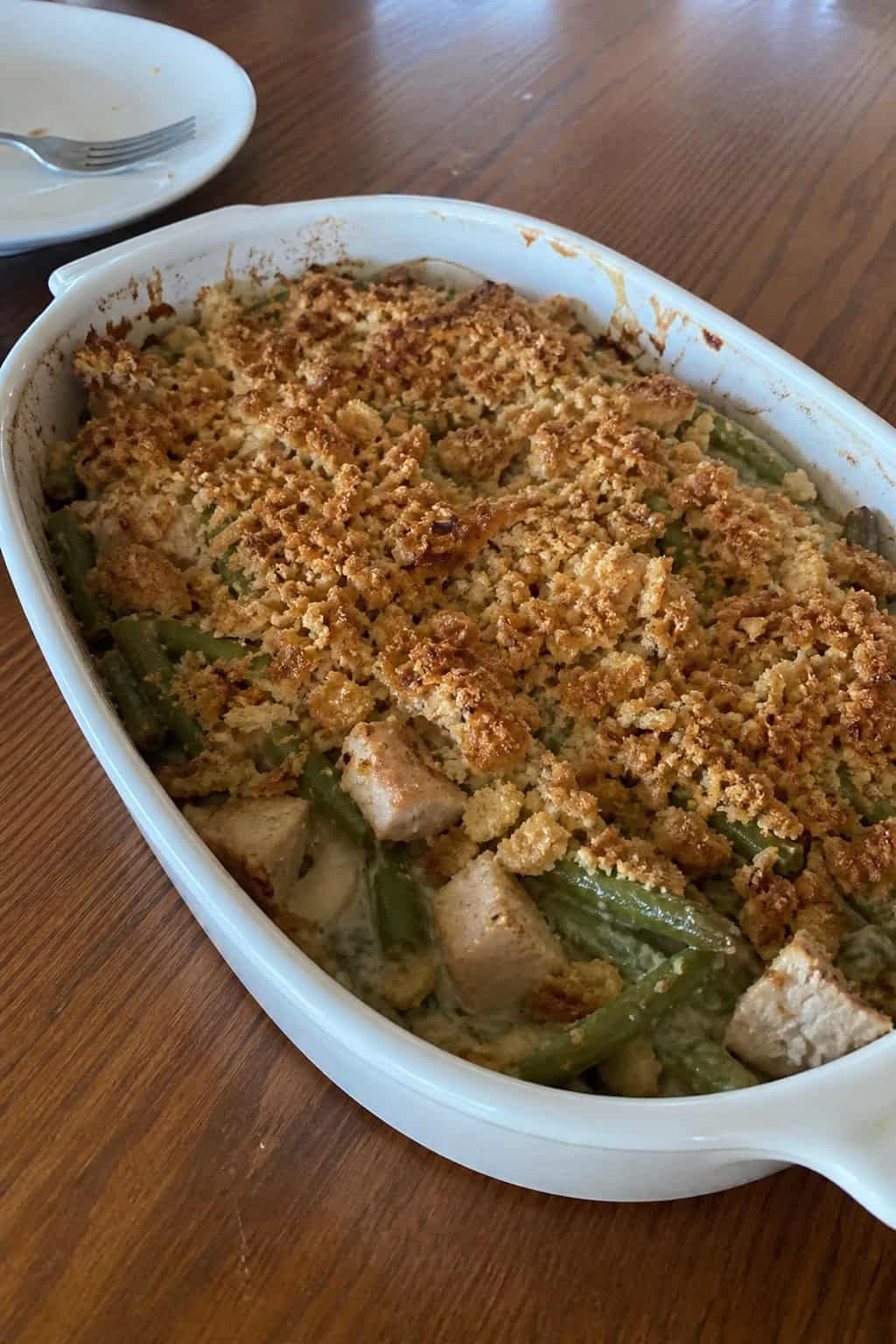 A baked green bean and turkey casserole in a white ceramic dish on a wooden table, with warm autumn-toned decorations in soft focus in the background.