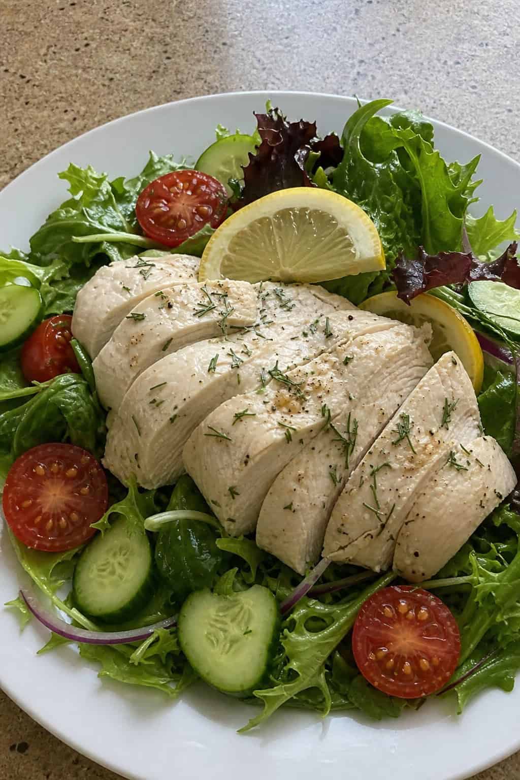 A plate of lemon-dill poached chicken breast slices served on a fresh green salad with cherry tomatoes and cucumber.