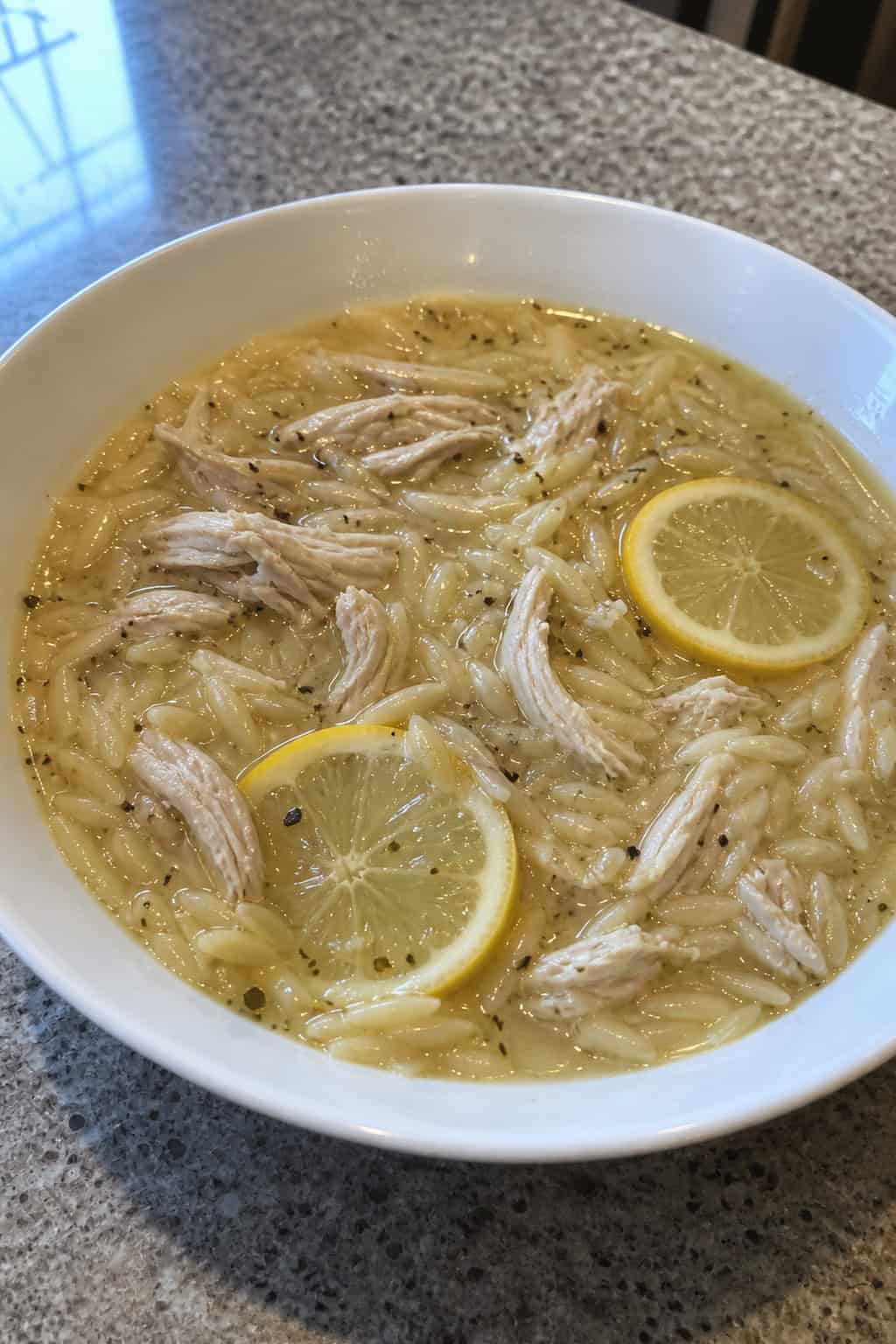 A ceramic bowl of golden chicken orzo soup with visible shredded chicken, tiny orzo pasta, wilted spinach, and thin lemon slices floating on top, surrounded by fresh lemons, garlic cloves, and parsley sprigs on a wooden table.