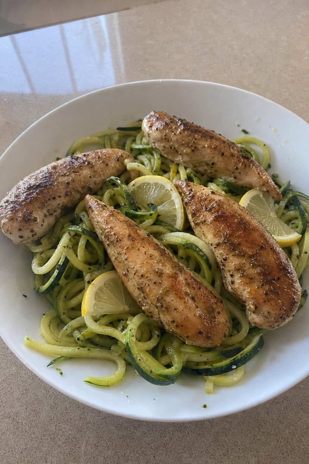 A plate of lemon-garlic chicken tenders served on zucchini noodles with lemon slices and herbs on a wooden table.