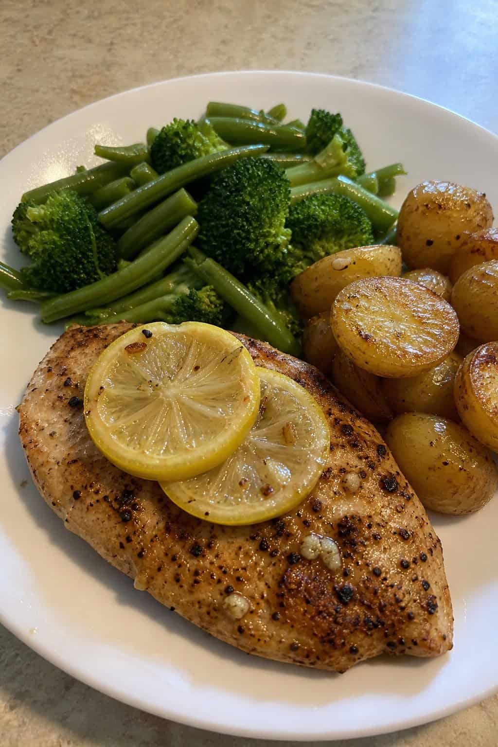 A plate with a lemon-garlic pan-seared chicken breast garnished with lemon slices and herbs, served with green vegetables and roasted potatoes on a wooden table.