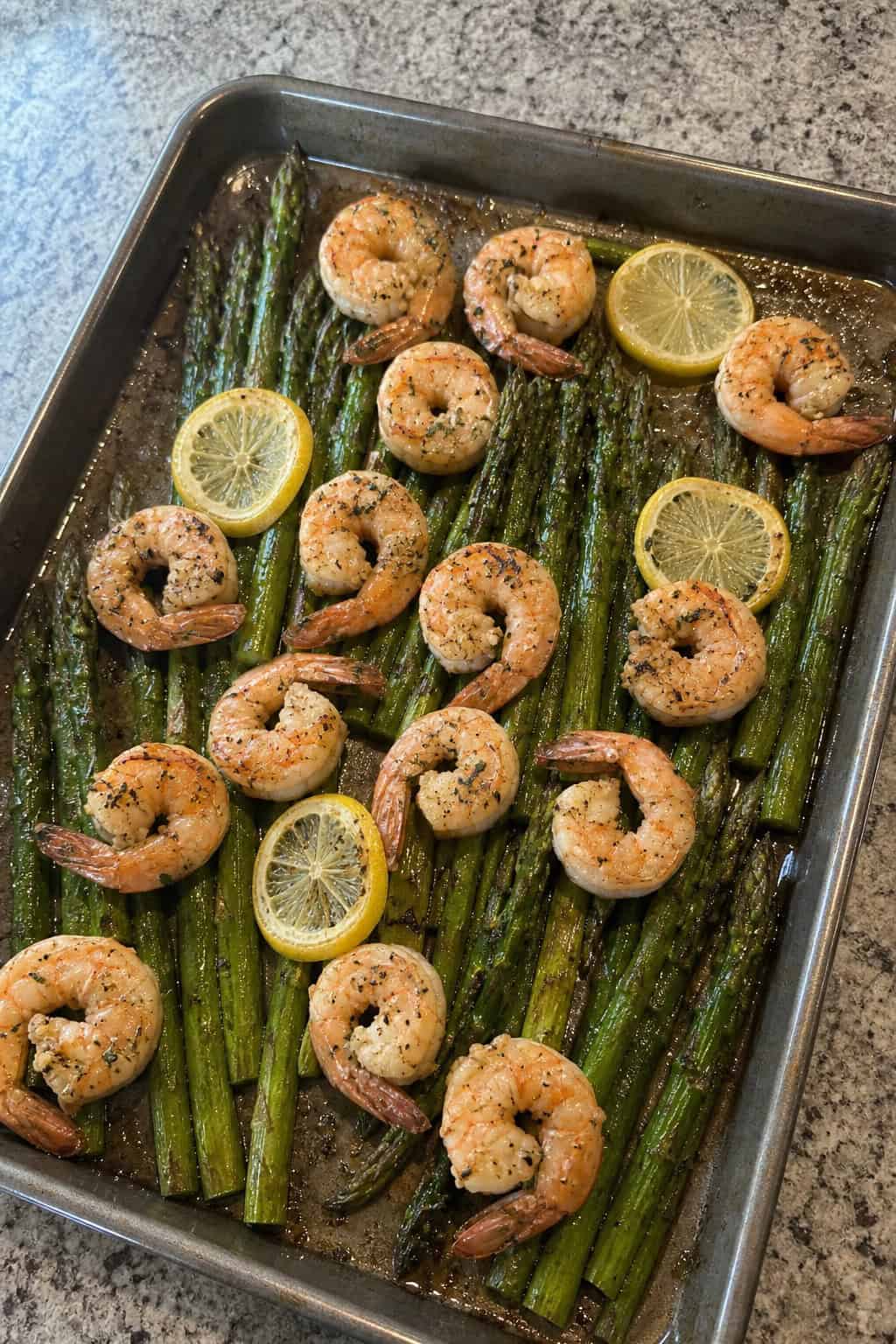 Plump pink shrimp and bright green asparagus spears roasted on a foil-lined sheet pan, garnished with thin lemon rounds and fresh parsley, set on a pale wooden surface.